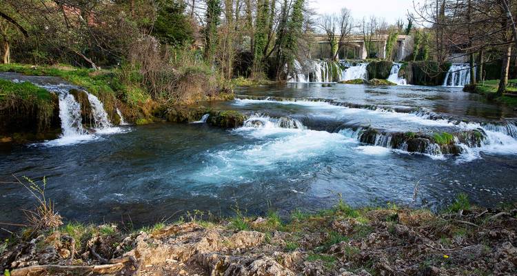 Lage trapsgewijze watervallen die over rotsen naar beneden stromen in een rustige rivier omzoomd met groen.