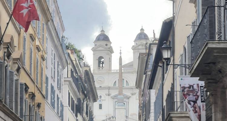 Estrecha calle romana que conduce a los Escalinata Española y a la iglesia de Trinità dei Monti con sus torres gemelas bajo un cielo dramático.