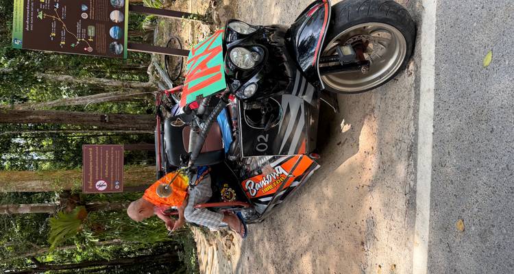 An older man sits on a custom three-wheeled motorbike parked beside a forest road.