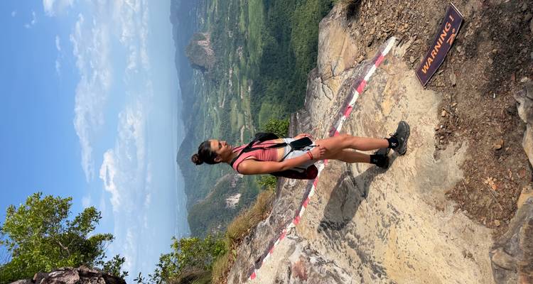A woman stands on a rocky cliff edge with sweeping views of green valleys and limestone hills below.