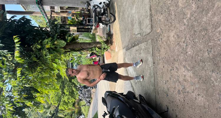 A shirtless man stands on a concrete driveway beside parked motorbikes in a tropical setting.
