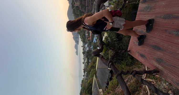A woman looks out over a coastal village and bay from a wooden viewpoint at dusk.