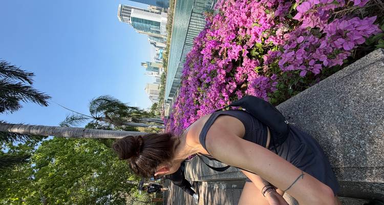 A woman sits on a park ledge next to bright magenta flowers overlooking a lake and Bangkok skyline on a sunny day.