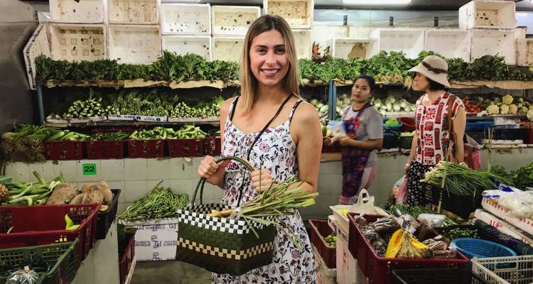 Market scene with woman holding basket of vegetables surrounded by fresh produce stalls.