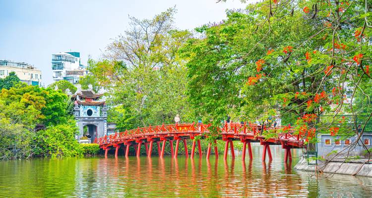 Puente de madera rojo brillante que se extiende a lo largo de un lago tranquilo hacia una pequeña puerta de pagoda, enmarcado por árboles frondosos.