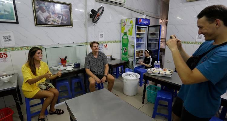 Grupo de viajeros riendo y tomando fotos mientras están sentados en pequeños banquitos en un restaurante vietnamita casual.