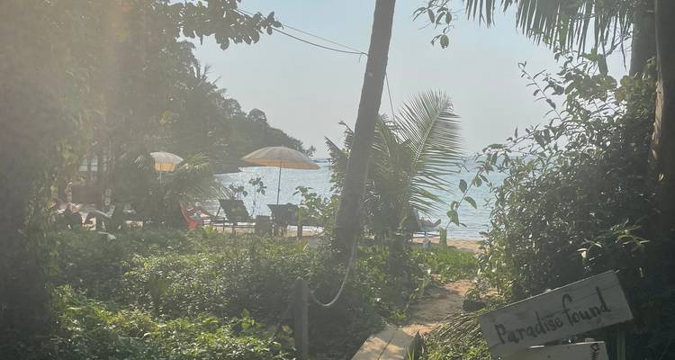 Backlit garden path leading to beach umbrellas and sea, with sign reading 'Paradise found'.