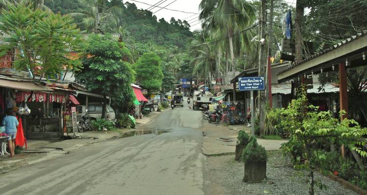 Rural Thai village road lined with small shops, greenery and power lines amidst forested hills.