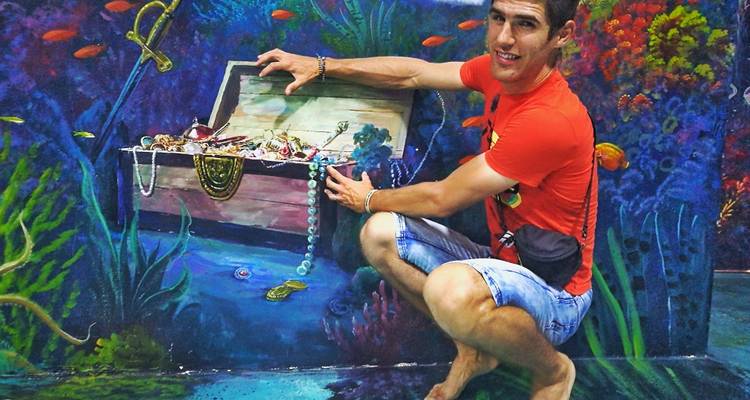 Man kneels beside a painted treasure chest overflowing with jewels in a vibrant underwater-themed illusion.