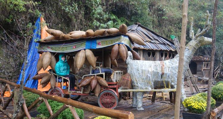 Eine Person in einer blaugrünen Regenjacke fährt einen rustikalen Wagen, der mit geflochtenen Körben verziert ist, neben einem Pappmaché-Tier in einem nebligen Bergdorf.