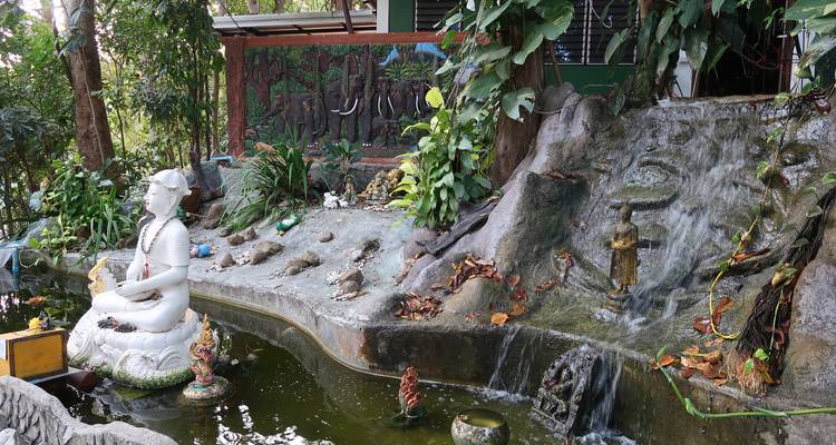 Üppiger Ziergarten mit einem weißen Meditationsstatue, Miniatur-Wasserfällen und tropischer Begrünung.