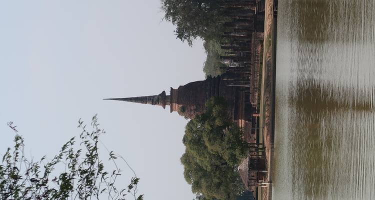 Backstein-Stupa mit einem schlanken Spitzturm, der neben einem reflektierenden See und einer Baumreihe unter blauem Himmel aufragt.