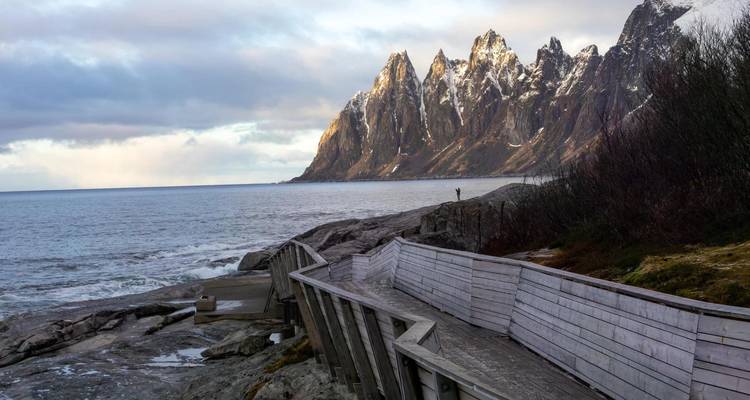 Une passerelle en bois épouse une côte rocheuse face à des pics dentelés saupoudrés de neige de l'autre côté de la mer.