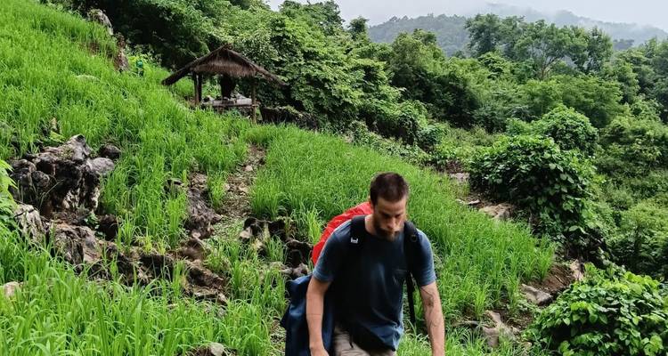 Een alleenstaande wandelaar met een rode rugzak loopt omhoog over een smal zandpad door weelderige groene hellingsrijstterrassen en junglevegetatie.