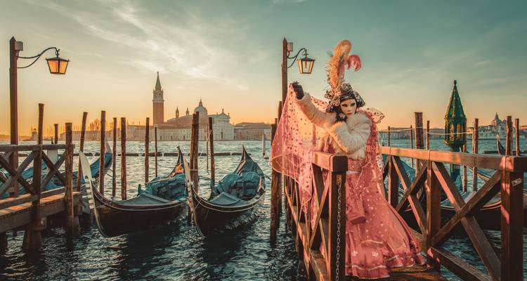 Eine kostümierte Figur posiert auf einem hölzernen Steg mit Gondeln und der Venezianischen Skyline bei Sonnenaufgang.