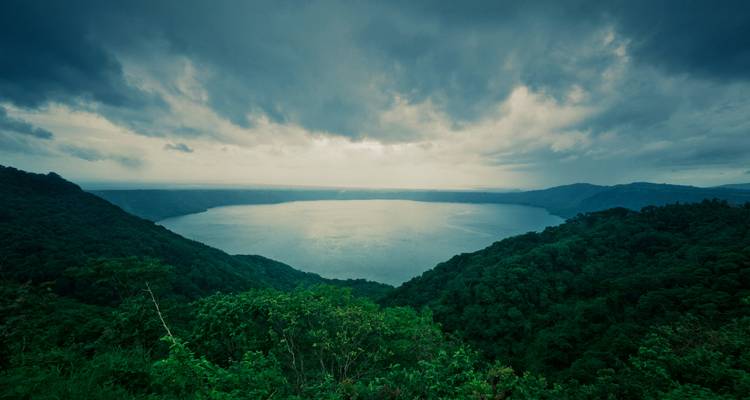 Moody panoramic of a circular crater lake surrounded by dense green forest under dramatic clouds.