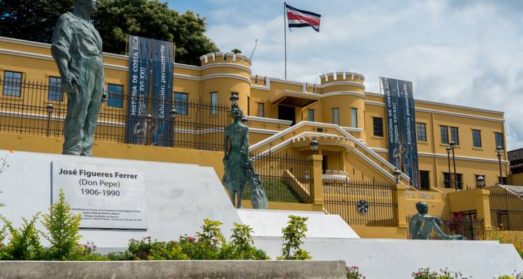 National Museum of Costa Rica with flag atop, flanked by bronze statues and banners.