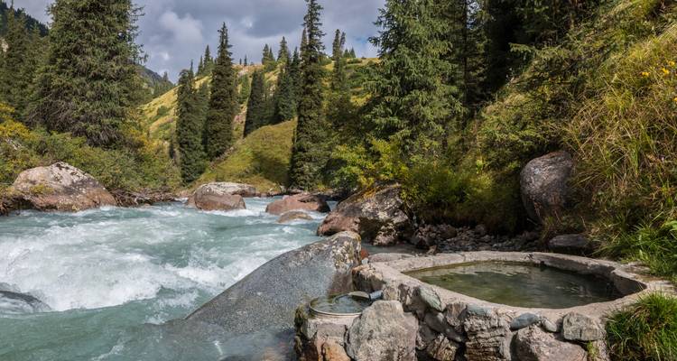 Bassin d'eau chaude naturelle situé à côté d'une rivière de montagne tumultueuse avec un arrière-plan de forêt de pins.