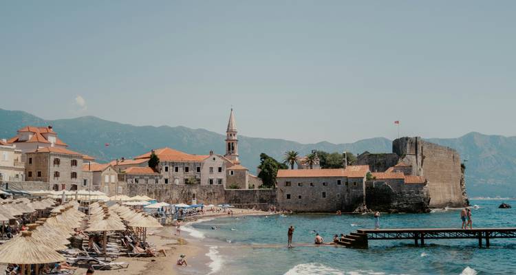 Plage de sable animée en bas de la vieille ville historique fortifiée de Budva avec des parasols, des baigneurs et les montagnes de l'Adriatique au loin.