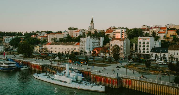 Promenade riveraine de Belgrade avec navires amarrés, bâtiments historiques et flèche d'église baignés dans la douce lumière du soir.