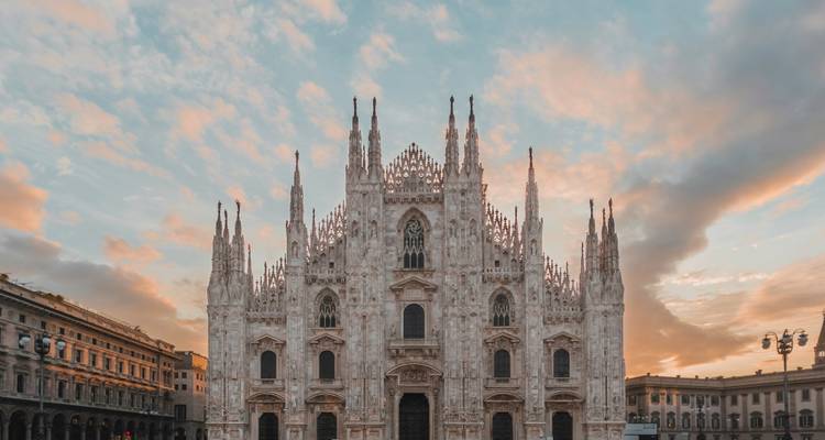 La majestueuse façade en marbre du Dôme de Milan s'élevant contre un ciel de coucher de soleil aux teintes pastel avec les bâtiments de la piazza environnante.
