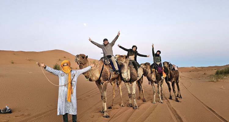 Camel caravan with riders raising their arms in celebration on a tranquil desert evening with a visible moon.
