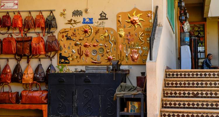 Leather goods and handcrafted curios on display outside a Moroccan workshop while a man sits nearby.