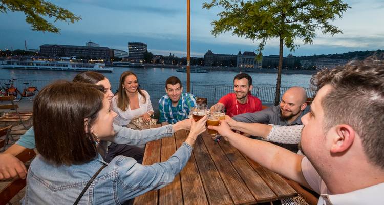 Des amis trinquent avec leurs verres de bière à une table au bord de la rivière surplombant le front de l'eau du Danube à Budapest.
