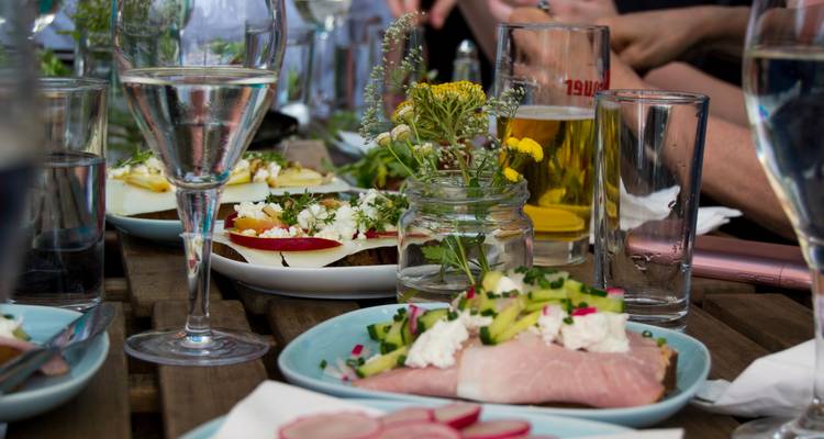 Gros plan d'une table extérieure rustique dressée avec des sandwichs ouverts, de la bière et de l'eau pétillante lors d'un repas décontracté.