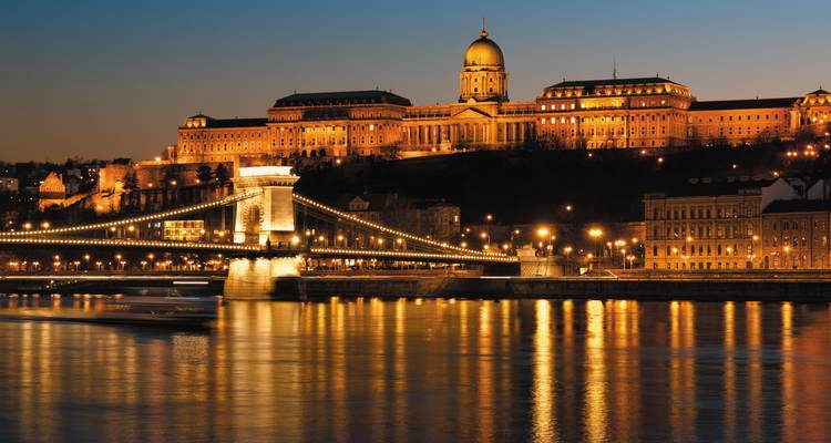 Vue nocturne du Pont des Chaînes de Budapest illuminé au-dessus du Danube avec le Château de Buda éclairé sur la colline à l'arrière-plan, reflété dans le fleuve calme.