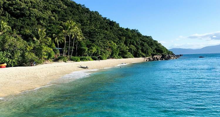 Turquoise water spoelt tegen een afgelegen tropisch strand met palmbomen en beboste heuvels erachter.