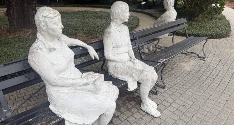 Des sculptures en ciment blanc de femmes assises sur des bancs de parc le long d'un chemin pavé de pierres.