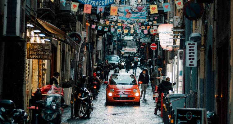 Busy narrow street in Naples with colorful banners, scooters, and a red Fiat moving through the crowd.