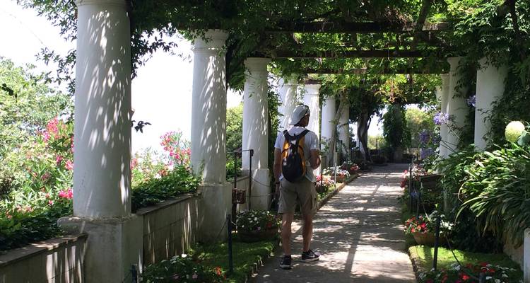 Tourist stands beneath a pergola of columns and vines in a lush Mediterranean garden.