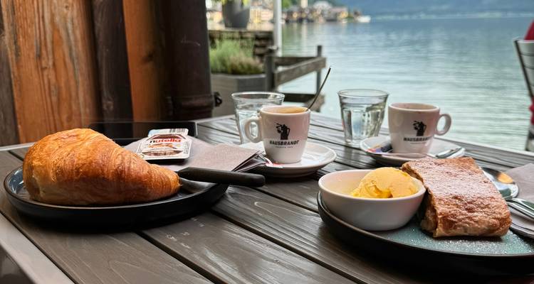 Table de petit-déjeuner dressée avec un croissant, une pâtisserie et un espresso surplombant un village pittoresque au bord du lac tranquille.