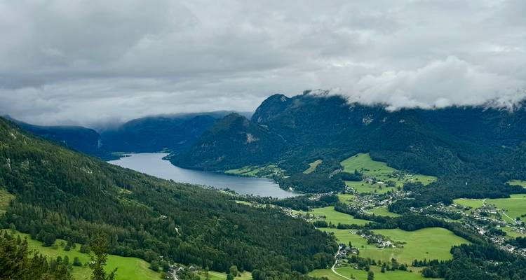 Vallée alpine verdoyante s'étendant vers un lac lointain sous des nuages ondulants et des versants boisés.