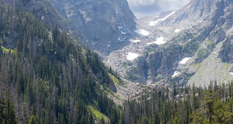 Steep, rugged Rocky Mountain valley with evergreen forests and lingering summer snow patches.