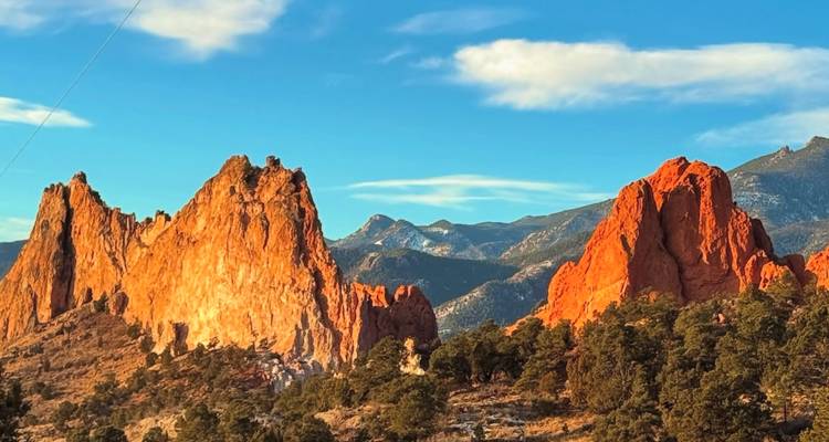 Sunlit red sandstone spires of Garden of the Gods rise dramatically against blue sky and distant mountains.