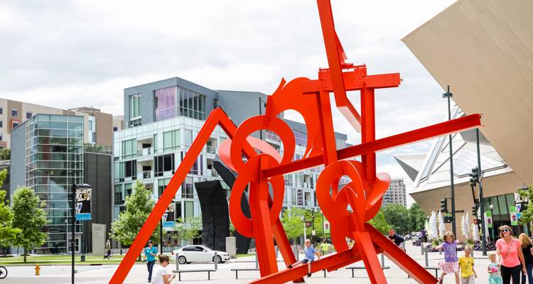 Bold red abstract sculpture stands in a modern downtown plaza with glass buildings and pedestrians.
