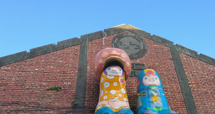 Estatuas de aspecto caricaturesco y coloridas frente a un almacén de ladrillo rojo bajo un cielo azul despejado.