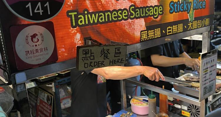 Puesto del mercado nocturno asando salchichas taiwanesas con un letrero colorido encima.