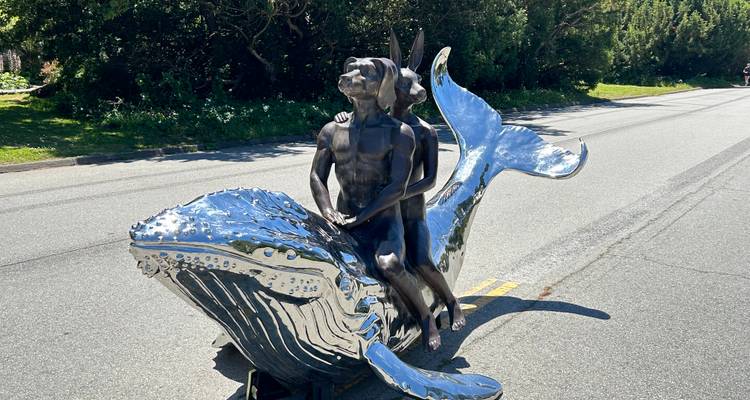 Reflective chrome sculpture of two figures riding a whale placed on a road in a park setting