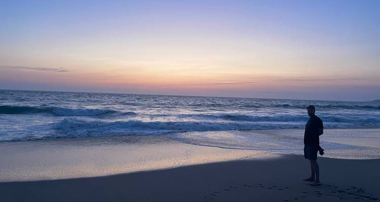 Silhouetted person standing on a sandy beach watching gentle waves at colorful sunset