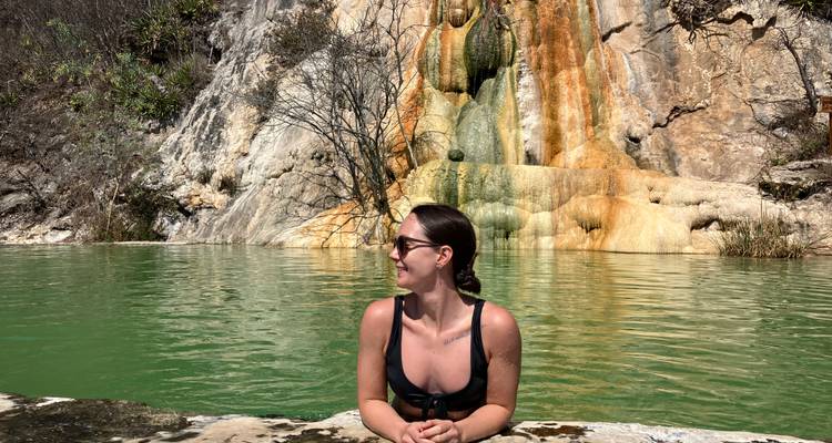 Woman relaxing in a natural mineral pool with colorful rock formations and waterfall backdrop at Hierve el Agua