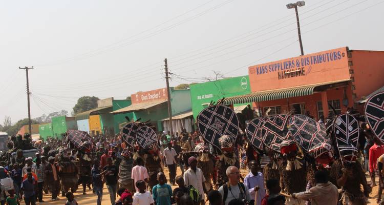 Défilé animé dans une ville avec des foules et des danseurs portant de grands boucliers aux motifs variés passant devant des devantures de magasins colorées.