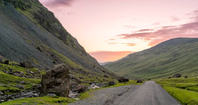 Narrow mountain road winding through steep green valley at colorful sunset.