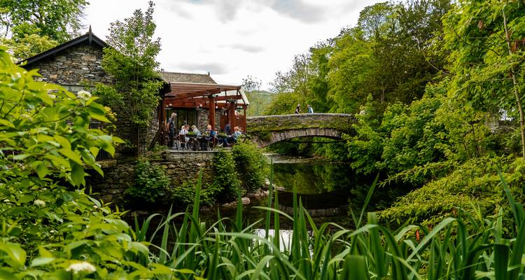 Leafy riverside cafe beside a small stone bridge in a lush woodland setting with diners enjoying the view.