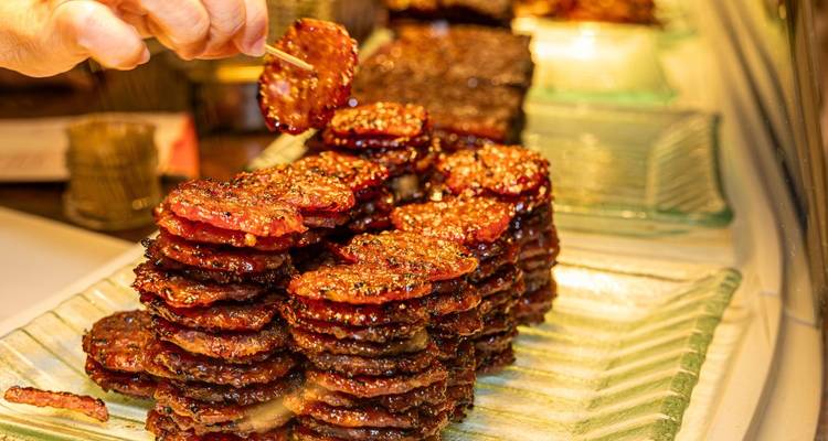 Stacks of glossy barbecued meat slices being prepared with a hand holding a skewer above them.