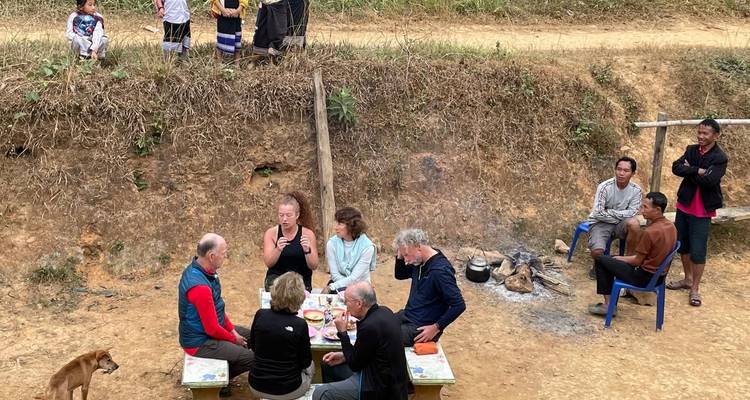 Small group of travellers share an outdoor meal on low benches in a rural village clearing.
