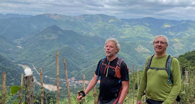 Two trekkers pause with trekking poles overlooking a wide river valley and green Laotian mountains.